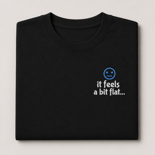 A folded black t-shirt on a light gray background, featuring a small blue sad face icon and the white text 'it feels a bit flat.