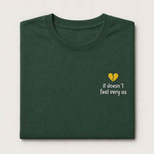 A folded dark green t-shirt with a small yellow broken-heart graphic and the white text 'it doesn't feel very us' embroidered on the left chest, displayed against a light off-white background.