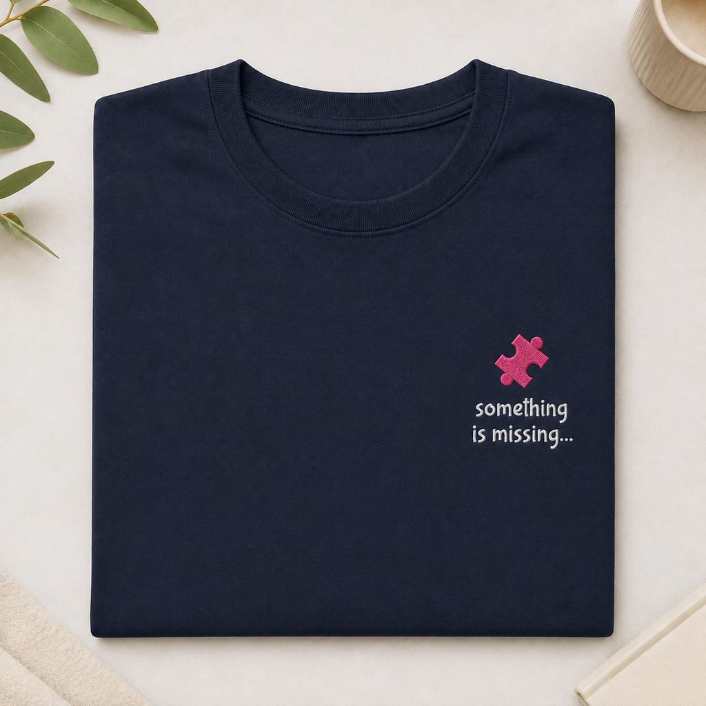 A folded navy blue t-shirt with a small pink puzzle piece graphic and the text 'something is missing.