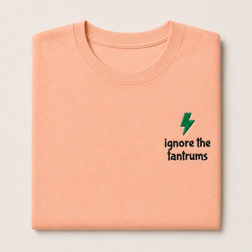 A folded peach-colored t-shirt with a small green lightning bolt graphic and the black text 'ignore the tantrums' printed on the left chest, set against a light gray background.