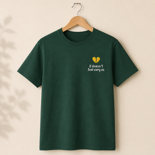 A dark green short-sleeve t-shirt hanging on a wooden hanger against a warm beige wall. The shirt features a small embroidered graphic on the left chest with a yellow broken-heart icon and the text 'it doesn't feel very us' in white.