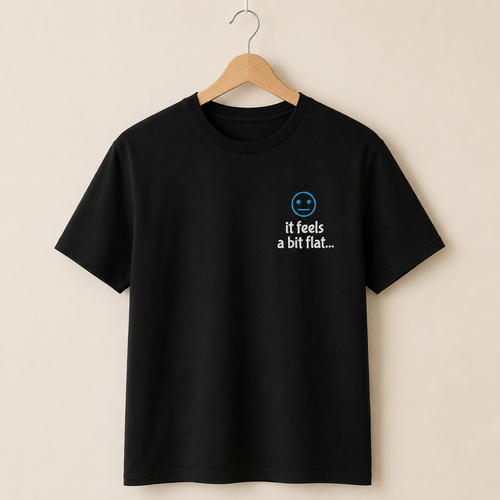 A black short-sleeve crewneck t-shirt hanging on a wooden hanger against a warm beige background. The shirt features a small graphic on the left chest with a blue smiley face and the text 'it feels a bit flat.