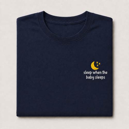 A folded navy blue t-shirt with a small embroidered graphic on the left chest showing a yellow crescent moon with 'z' symbols and the white text 'sleep when the baby sleeps', displayed on a light beige background.