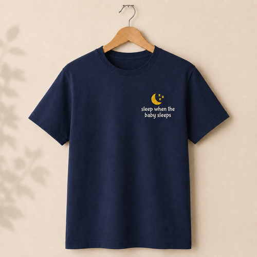 A navy blue short-sleeve t-shirt hanging on a wooden hanger against a warm beige background. The shirt features a small embroidered graphic on the left chest with a yellow crescent moon, white stars, and the text 'sleep when the baby sleeps' in white.