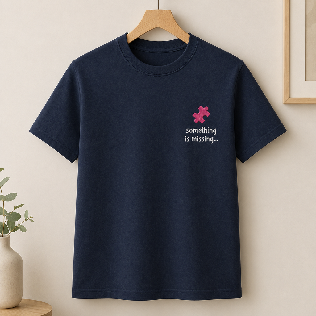 A navy blue short-sleeve crewneck t-shirt hanging on a wooden hanger against a warm beige wall. The shirt features a small pink puzzle piece graphic and the white text 'something is missing.