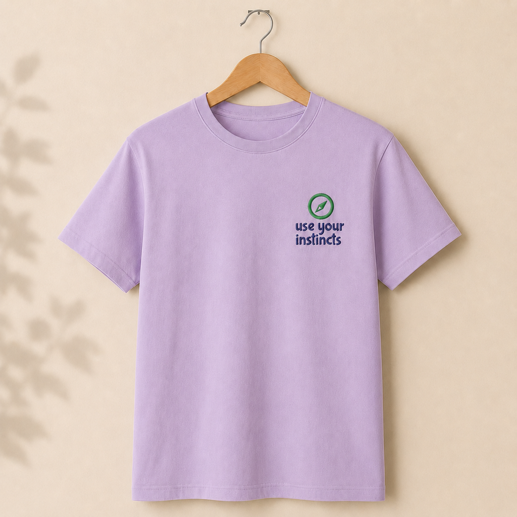 A light purple short-sleeve t-shirt hanging on a wooden hanger against a warm beige background. The shirt features a small graphic on the left chest with a green circle and checkmark icon above the navy blue text 'use your instincts'.