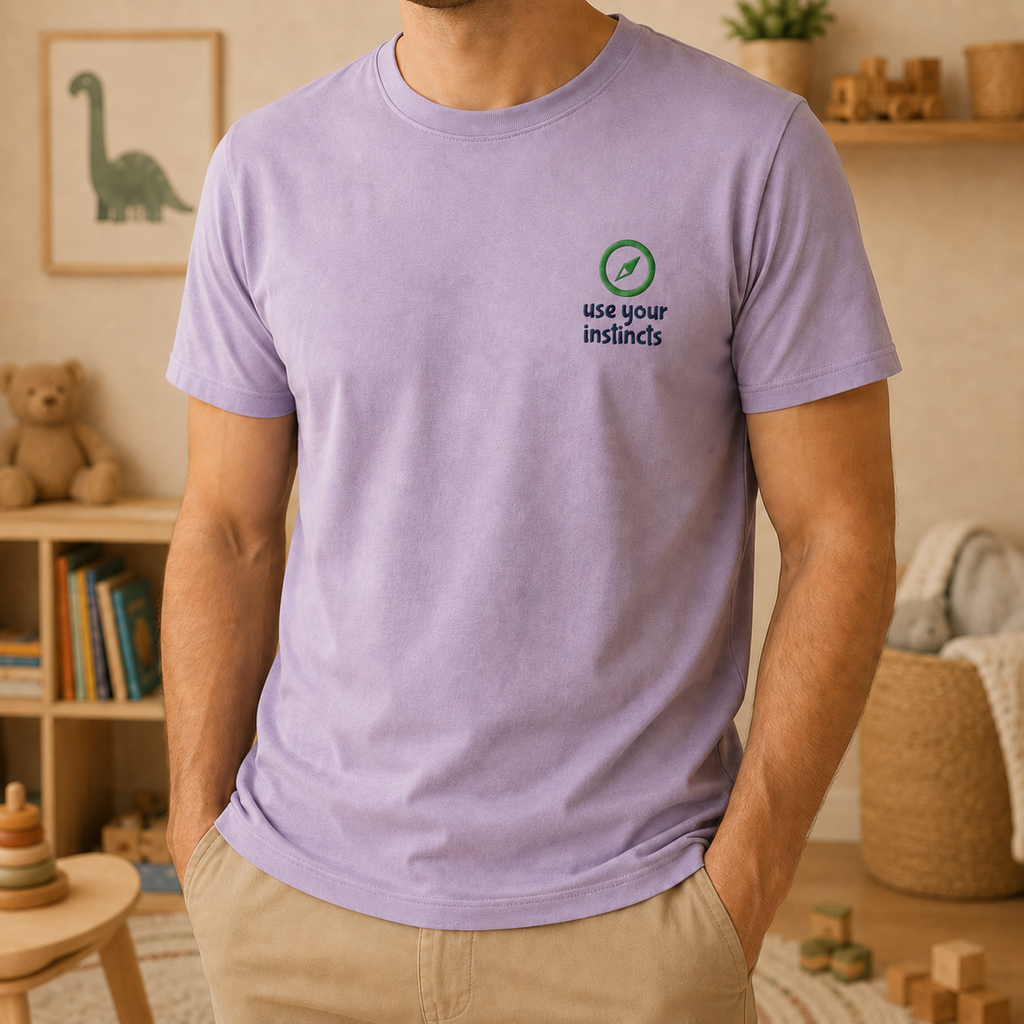 A man wearing a light purple crewneck t-shirt with a small graphic on the left chest that reads 'use your instincts' with a green and black symbol. The background shows a cozy, warm-toned child's room with toys, shelves, and a framed dinosaur picture.