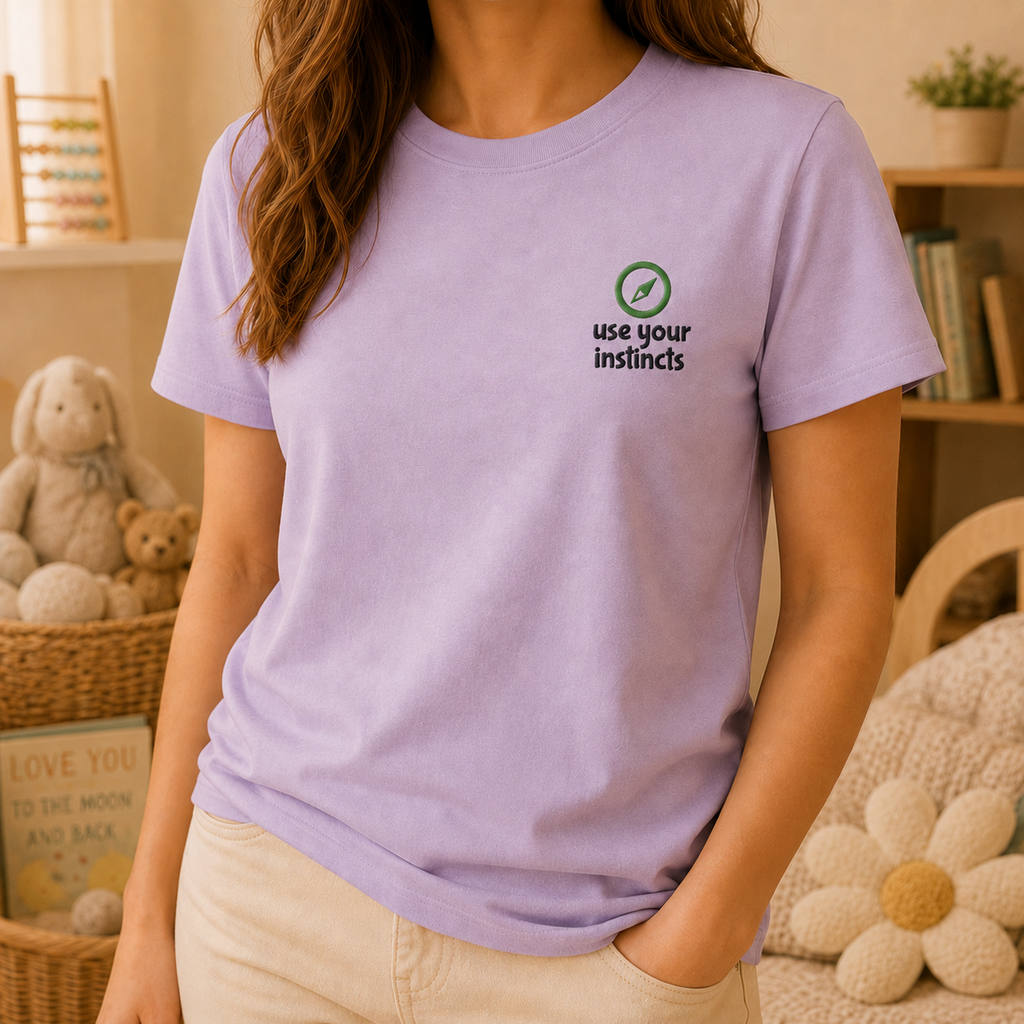 A woman wearing a light purple crewneck t-shirt with a small green and black graphic on the left chest that reads 'use your instincts' with a leaf icon. The background shows a cozy, warm-toned room with stuffed animals and a bookshelf.