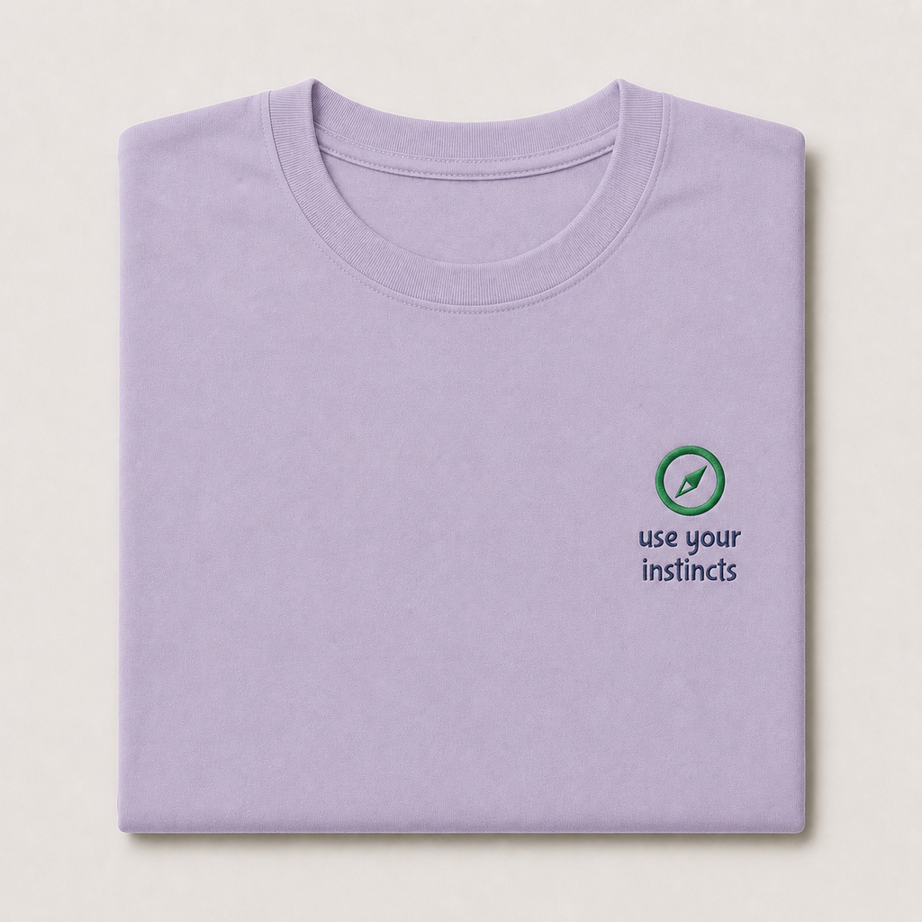 A folded light purple crewneck t-shirt on a light beige background, featuring a small green compass graphic and the text 'use your instincts' printed on the left chest.