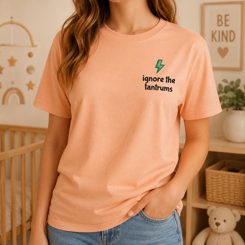A woman wearing a peach-colored short-sleeve t-shirt with a small graphic on the left chest that reads 'ignore the tantrums' in black text with a green lightning bolt above it. The background shows a softly lit nursery or bedroom setting with a crib, wall decorations, and a teddy bear.